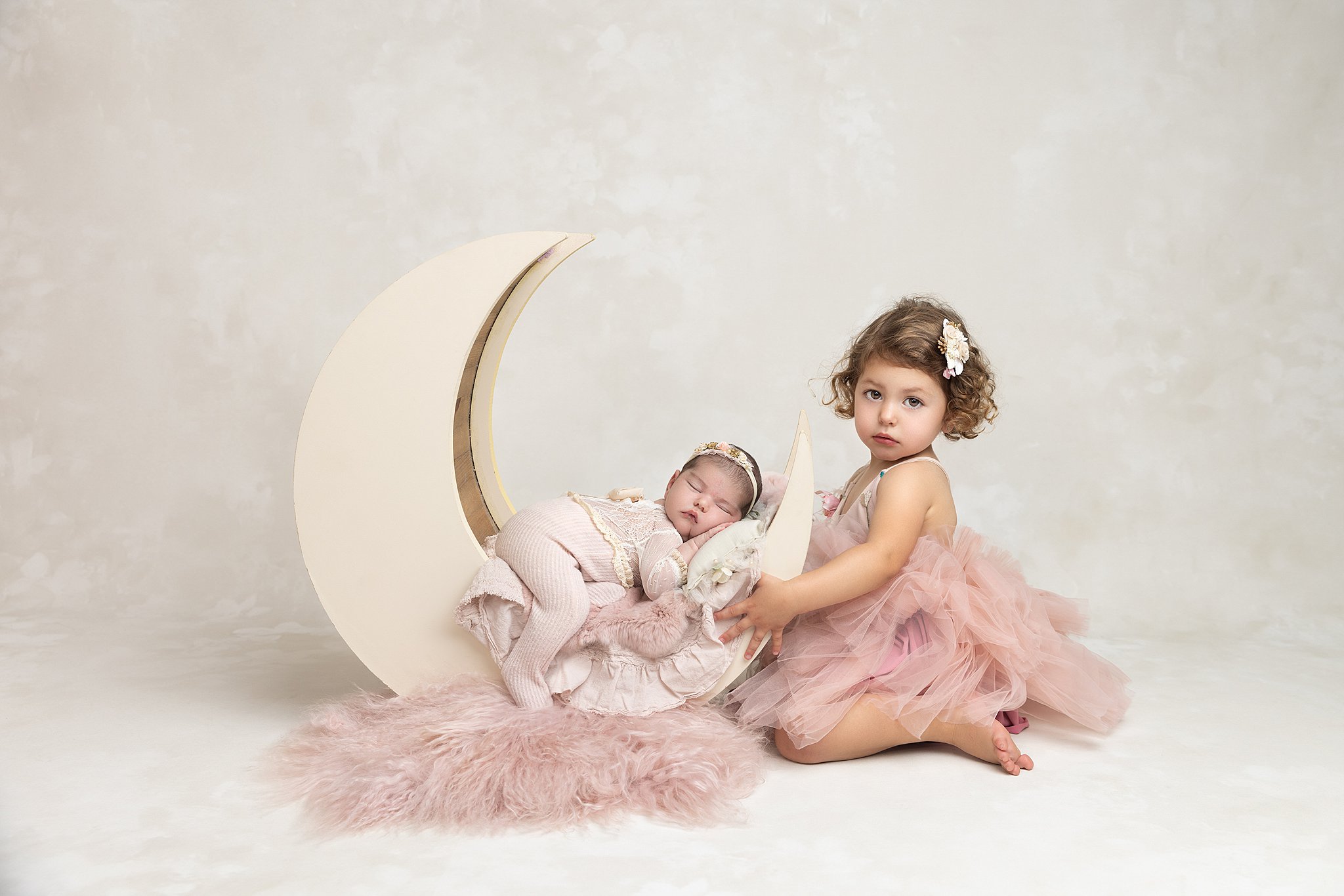 A toddler girl in a pink tutu kneels in a pink tutu with her sleeping baby sister in a moon shaped cradle