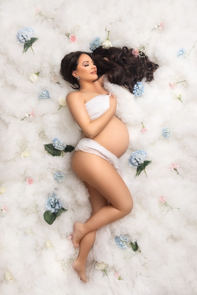mom lays in a bed of white tulle with pink and blue flowers to highlight that she is expecting twins during her maternity in nj