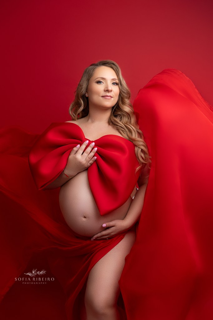 mom smiles to camera while draped in a red bow and fabric with a perth amboy maternity photographer