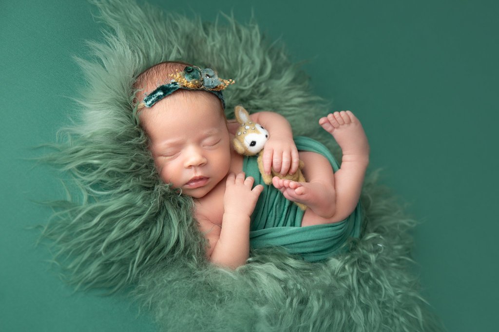 A newborn baby sleeps in a green swaddle with a felt deer toy and matching green headband thanks to postpartum place chatham