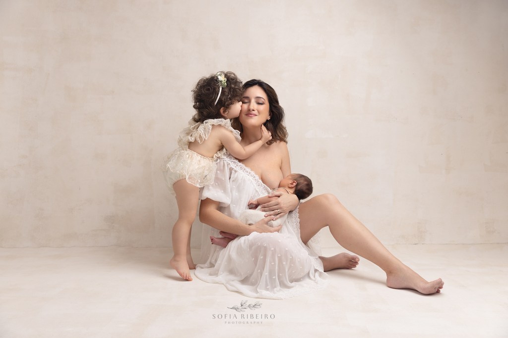 a mom sits with her newborn baby while getting a kiss from big sister during a session with a red bank nj newborn photographer