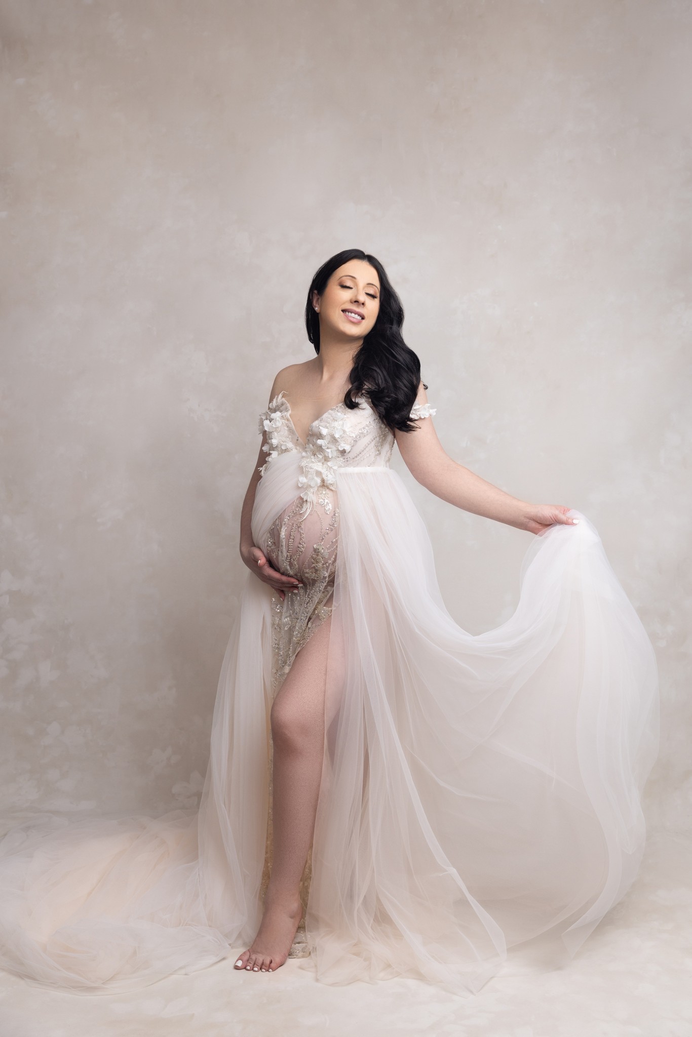 A smiling and joyful mother to be in an extravagant white maternity gown stands in a studio