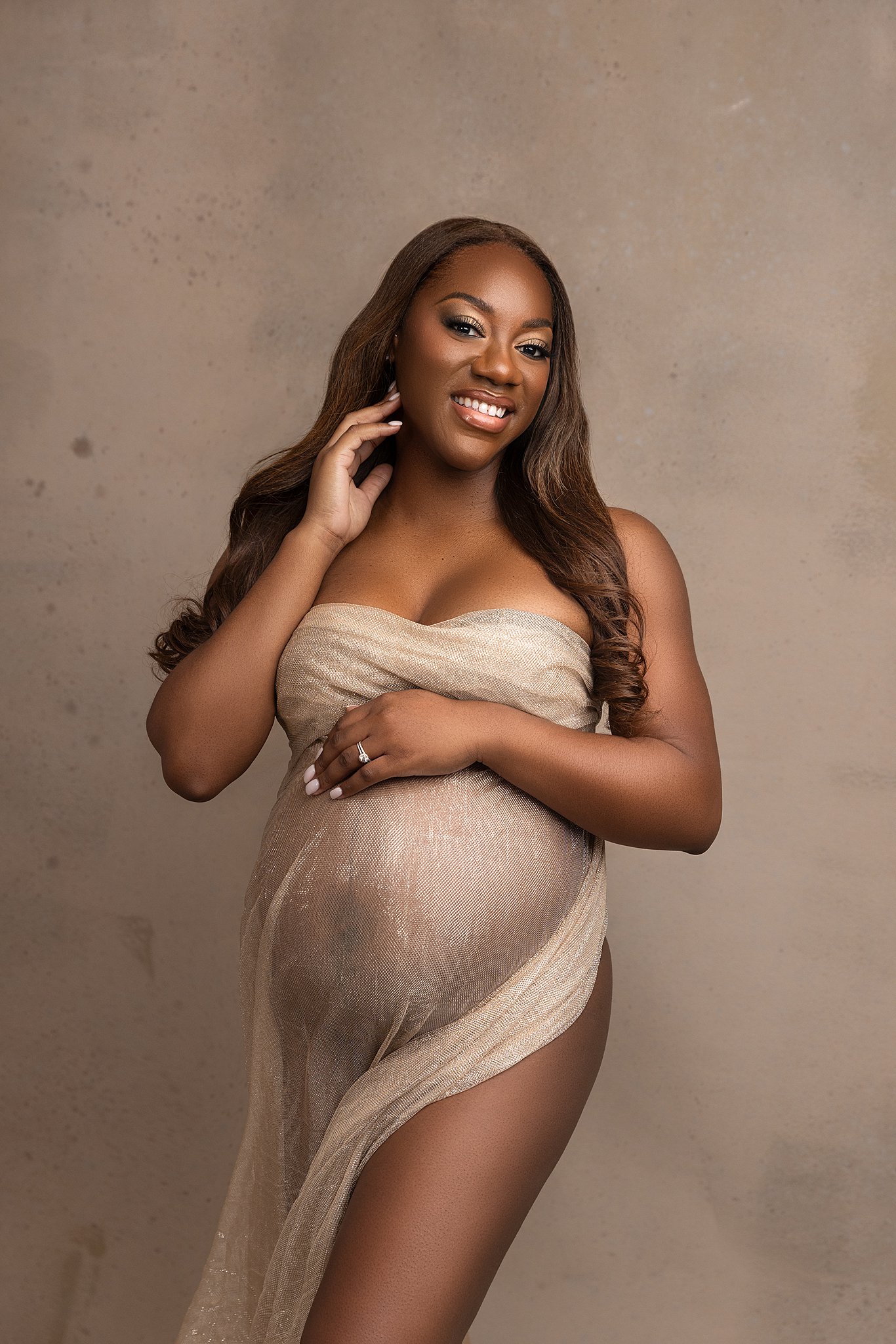 A pregnant woman wrapped in a sheer fabric smiles while brushing her hair back in a studio