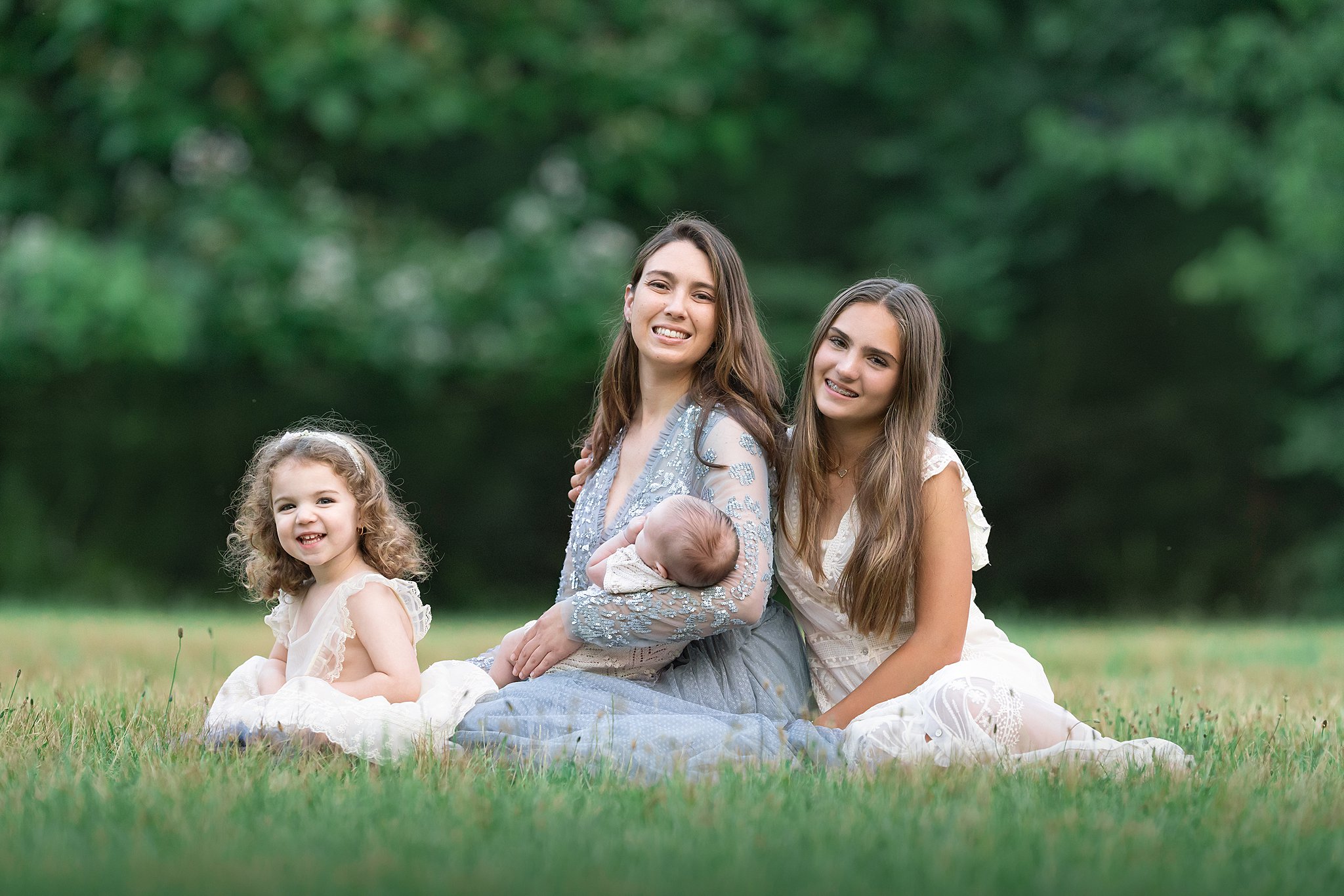 A happy mother sits in a park lawn holding her newborn baby with her teenage and toddler daughters in white dresses after visiting hoboken pediatricians