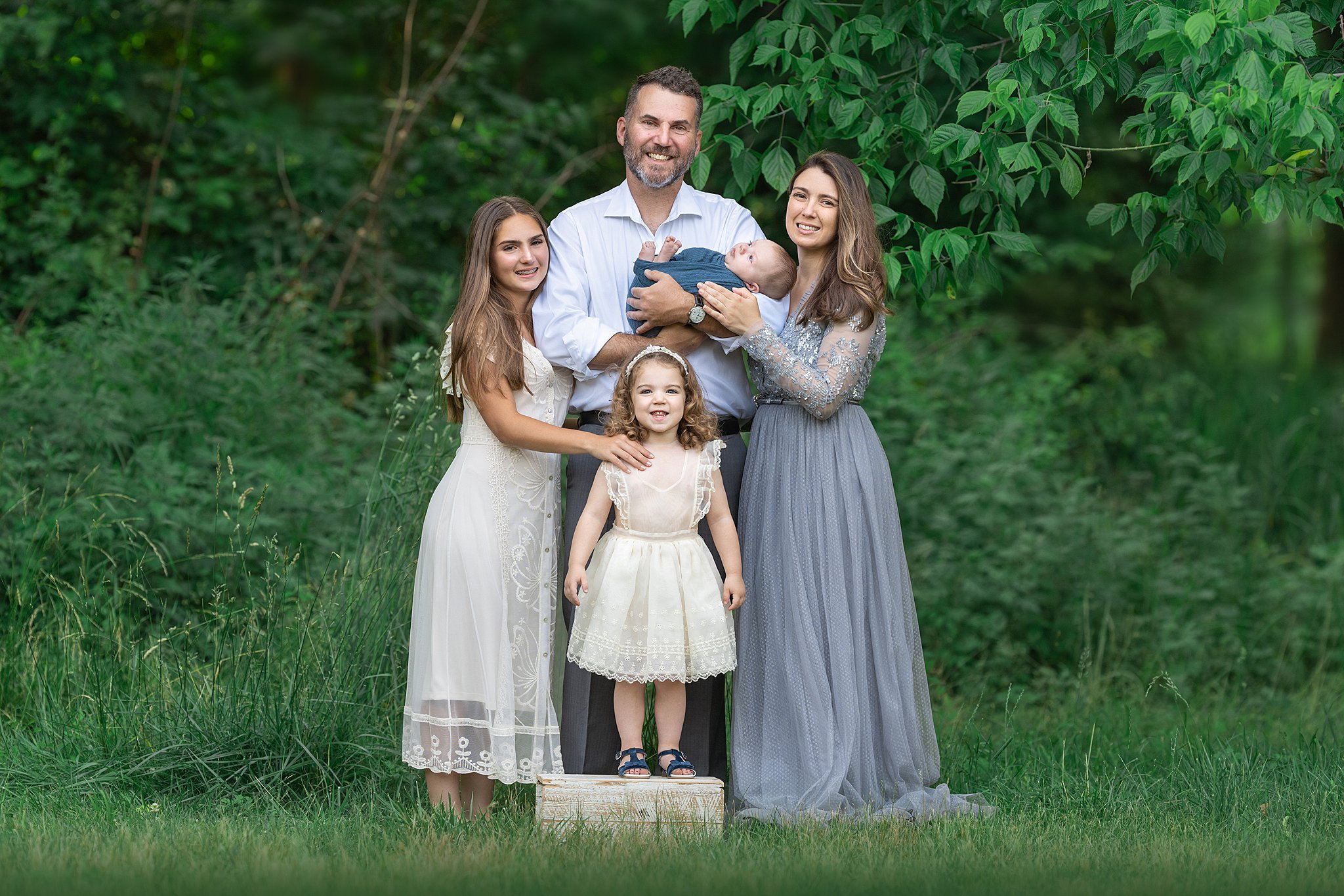 A happy family of 5 stand on the edge of a forest smiling and hugging