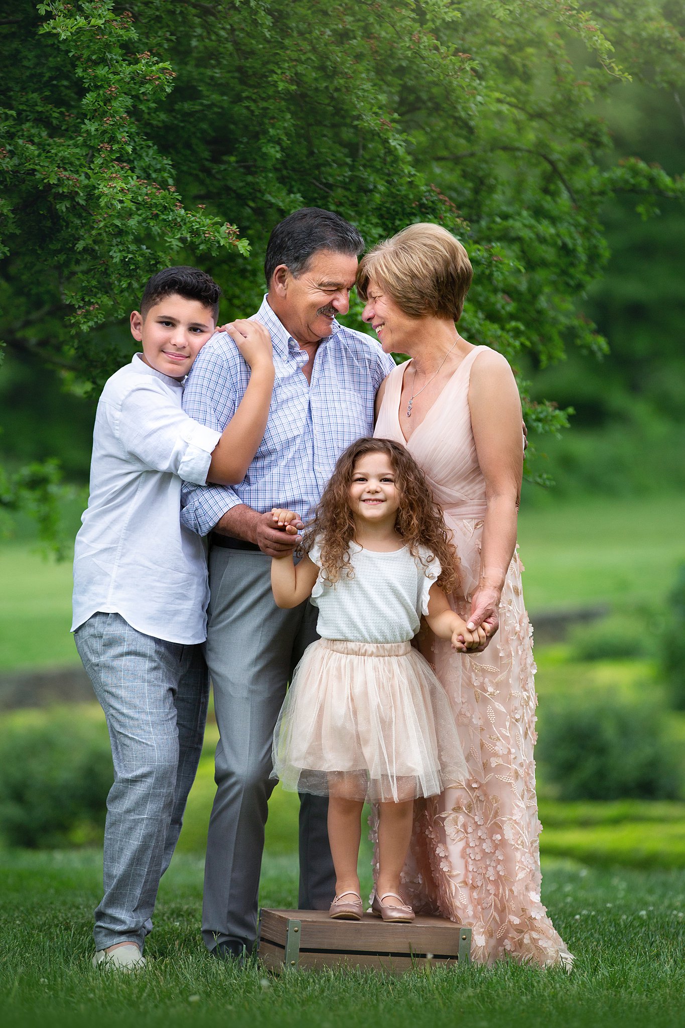 Happy parents stand in a park lawn touching foreheads and holding hands with their toddler daughter and teen son before visiting ice cream shops in nj