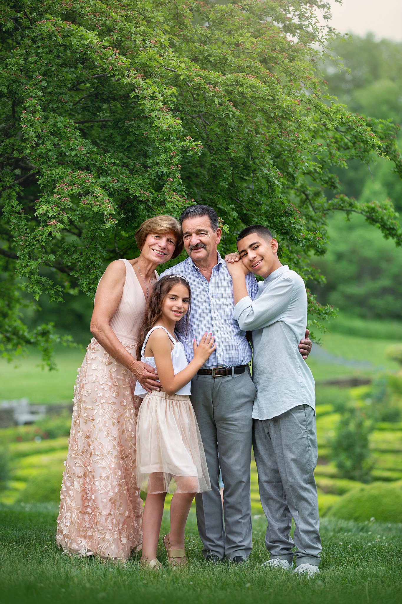 Happy parents stand under a tree in a garden hugging their young son and daughter before visiting ice cream shops in nj