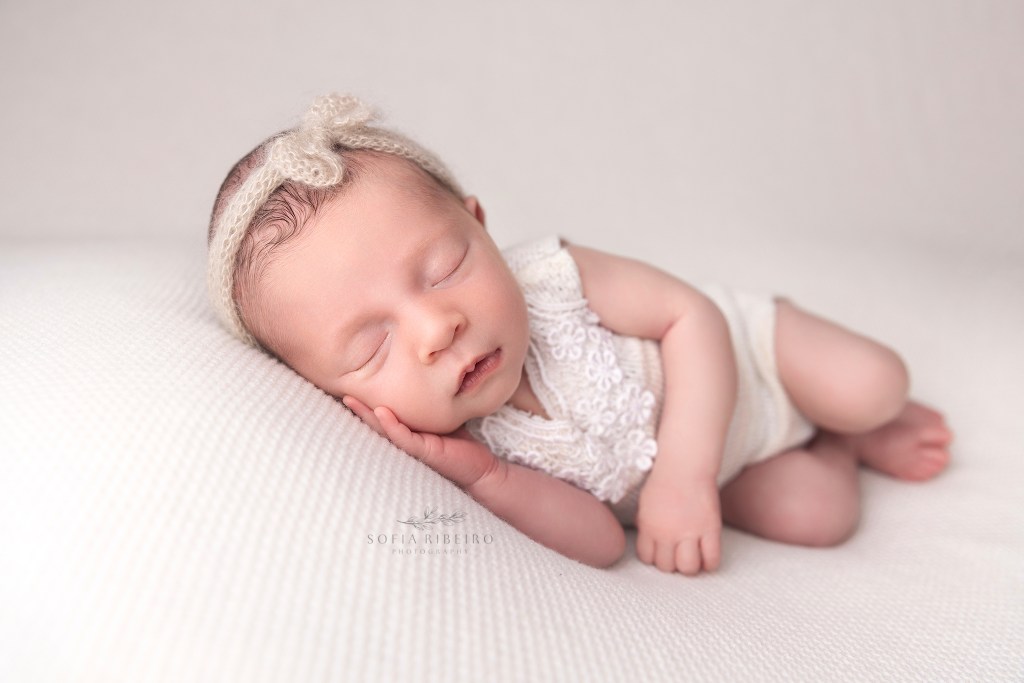 baby girl is dressed in a white onesie with a bow headband, posed on white fabric during her newborn photos