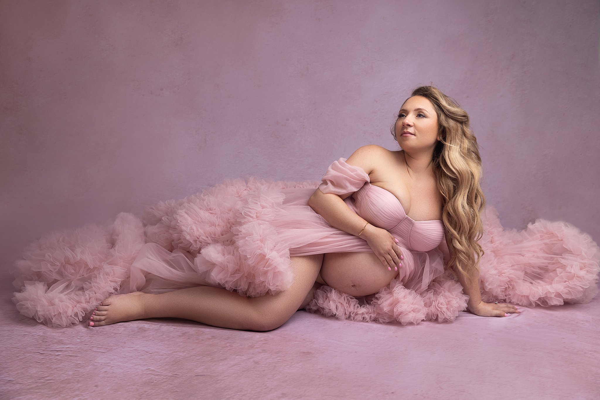 A mother to be in a flowing pink maternity gown smiles while laying across a matching studio floor after visiting on the side spa