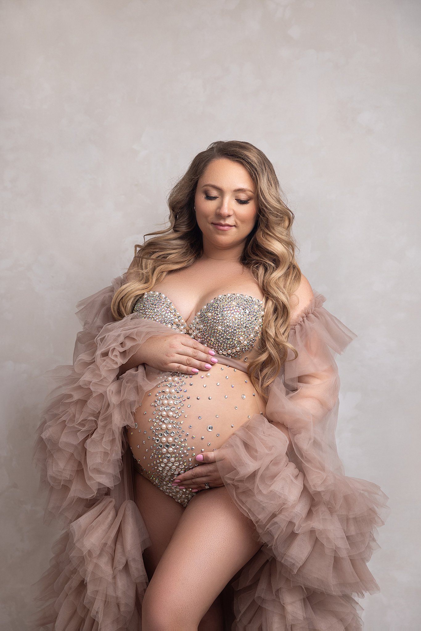 A mother to be smiles while holding her bump in a pearl covered maternity one piece with tule sleeves after visiting on the side spa