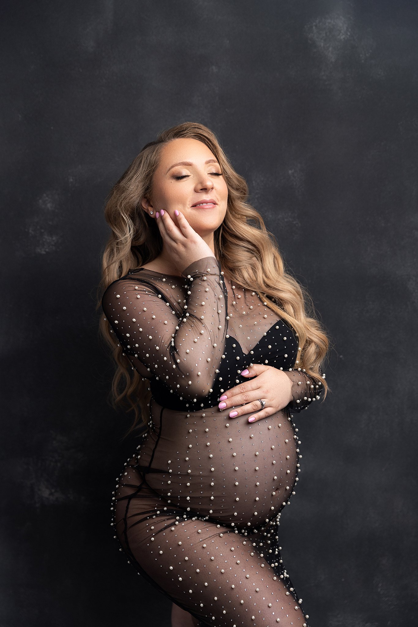 A happy pregnant woman holds her chin while standing in a sheer and pearl covered maternity gown