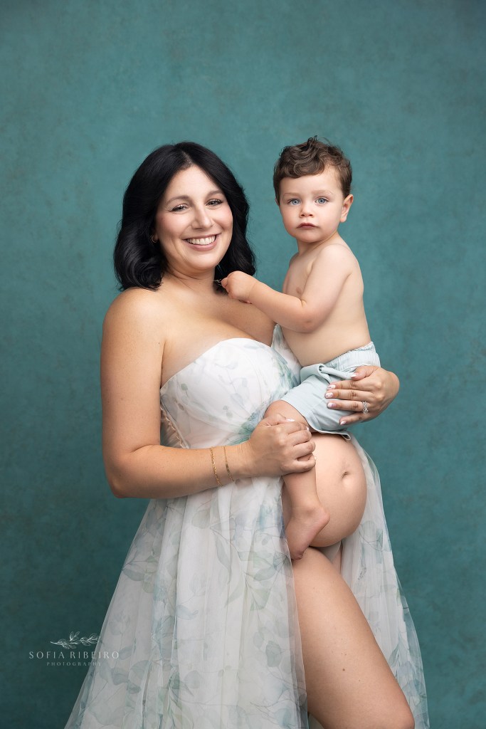 mom and her little boy pose together showing her belly against a teal backdrop during her maternity session in warren nj