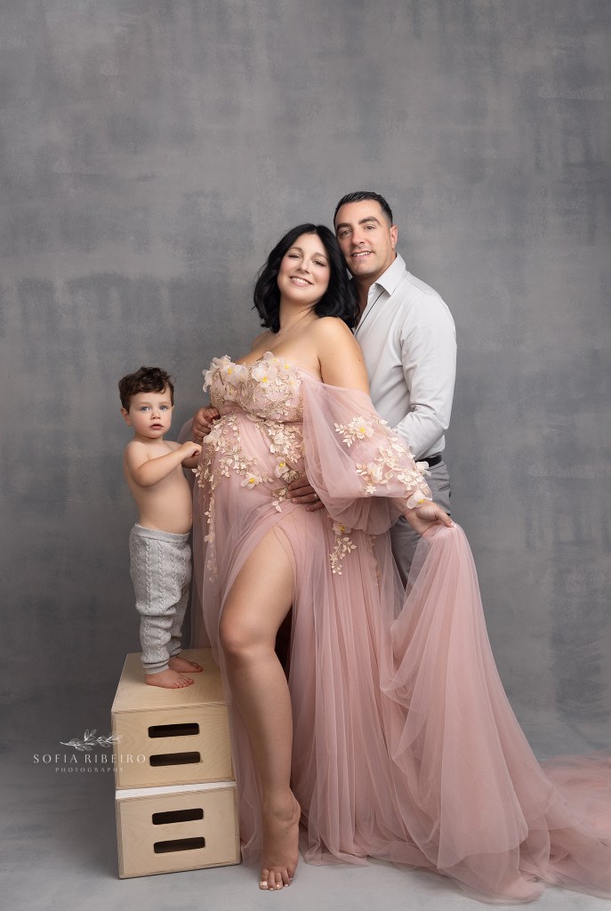 mom to be poses in a pink tulle gown with her family, smiling to camera in anticipation of a new baby in warren nj