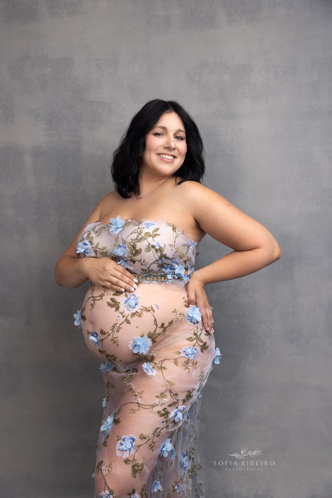mom smiles to camera, showing her bump in a sheer floral gown
