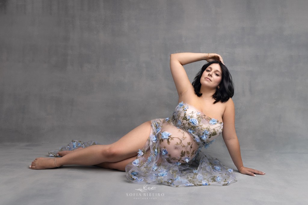 mom poses on the floor in a sheer blue floral applique fabric, highlighting her baby bump