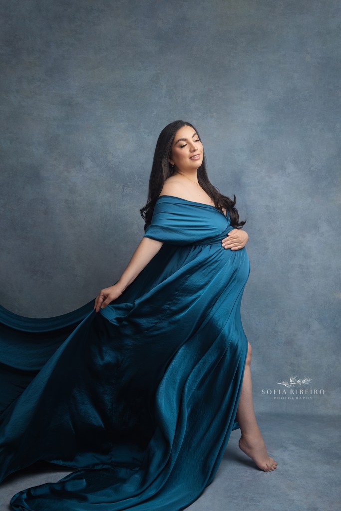 expectant mom holds the train of her flowing billowing dress with a fan blowing her hair and fabric back for a maternity portrait