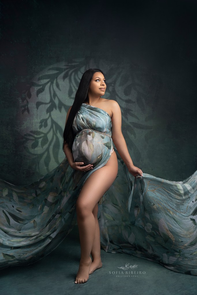 mom looks to the light as she holds a flowing printed fabric highlighting her pregnant belly