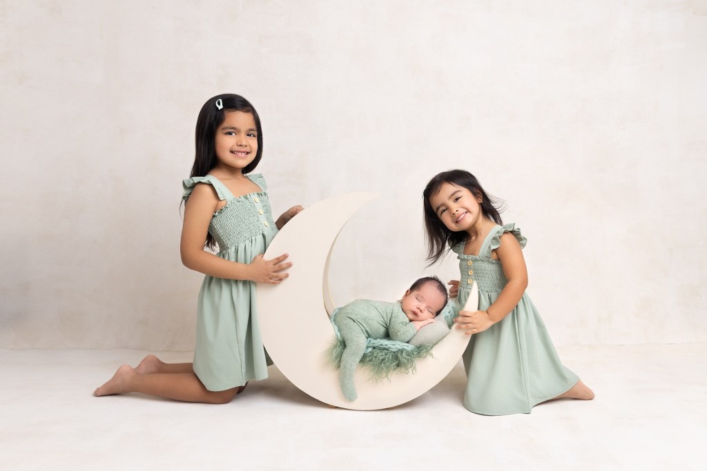 Happy toddler sisters in matching green dresses kneel in a studio with their newborn sibling sleeping in a moon crib after visiting madison nj pediatricians