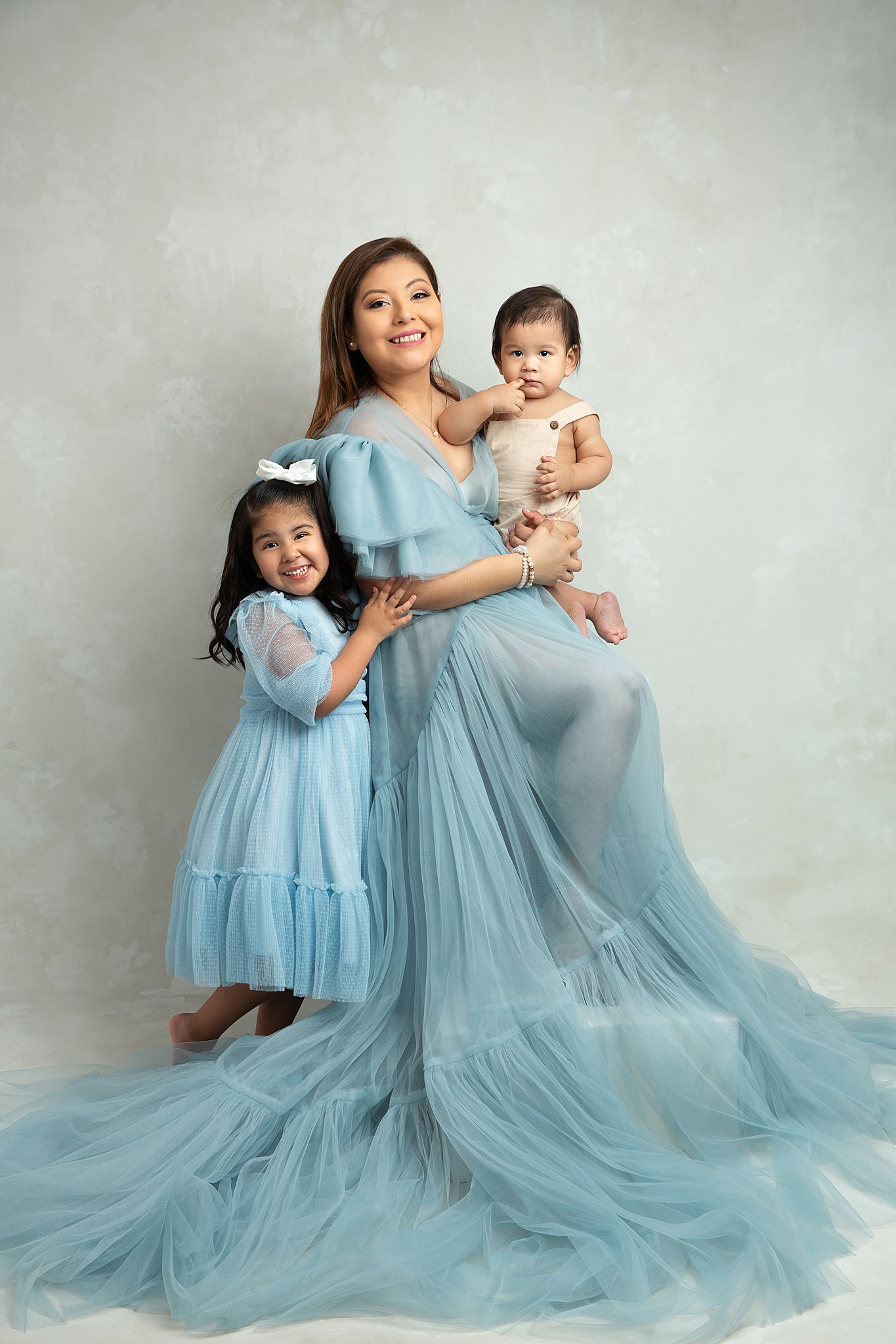 A happy mom in a long flowing dress sits in a studio smiling with her toddler daughter and infant son hugging her