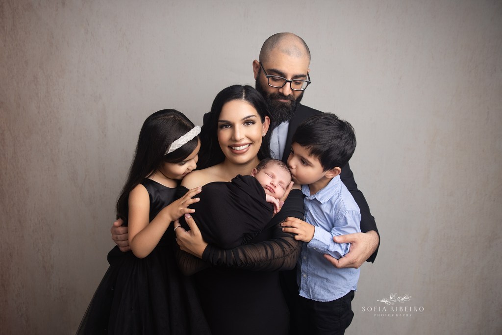 an adorable family photo of everyone looking at their new baby brother, all wearing black and blue against a light backdrop