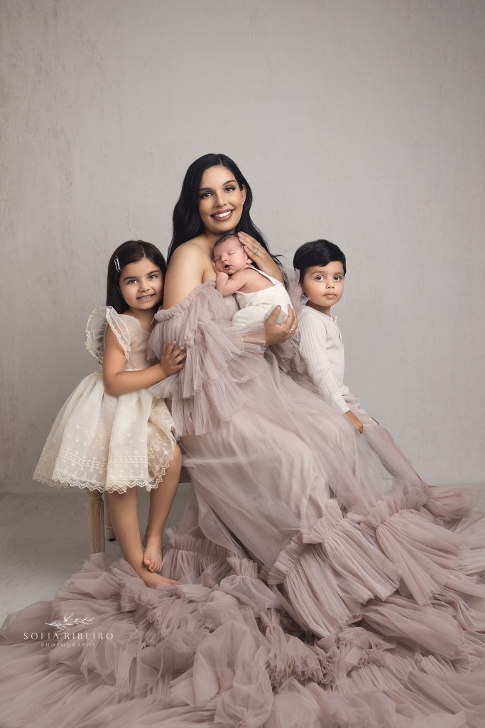mom dressed in tulle beige dress with all three of her children, against a cream backdrop for newborn phgotos