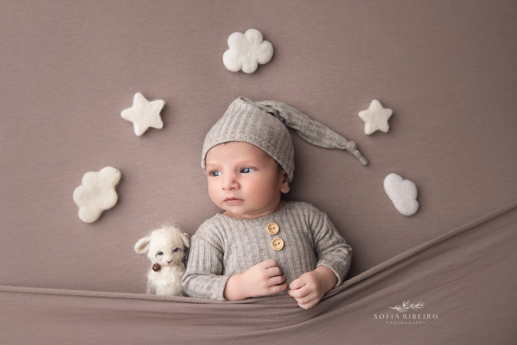 baby poses in a sleepy cap surrounded by clouds and stars over his head and a lamb stuffy