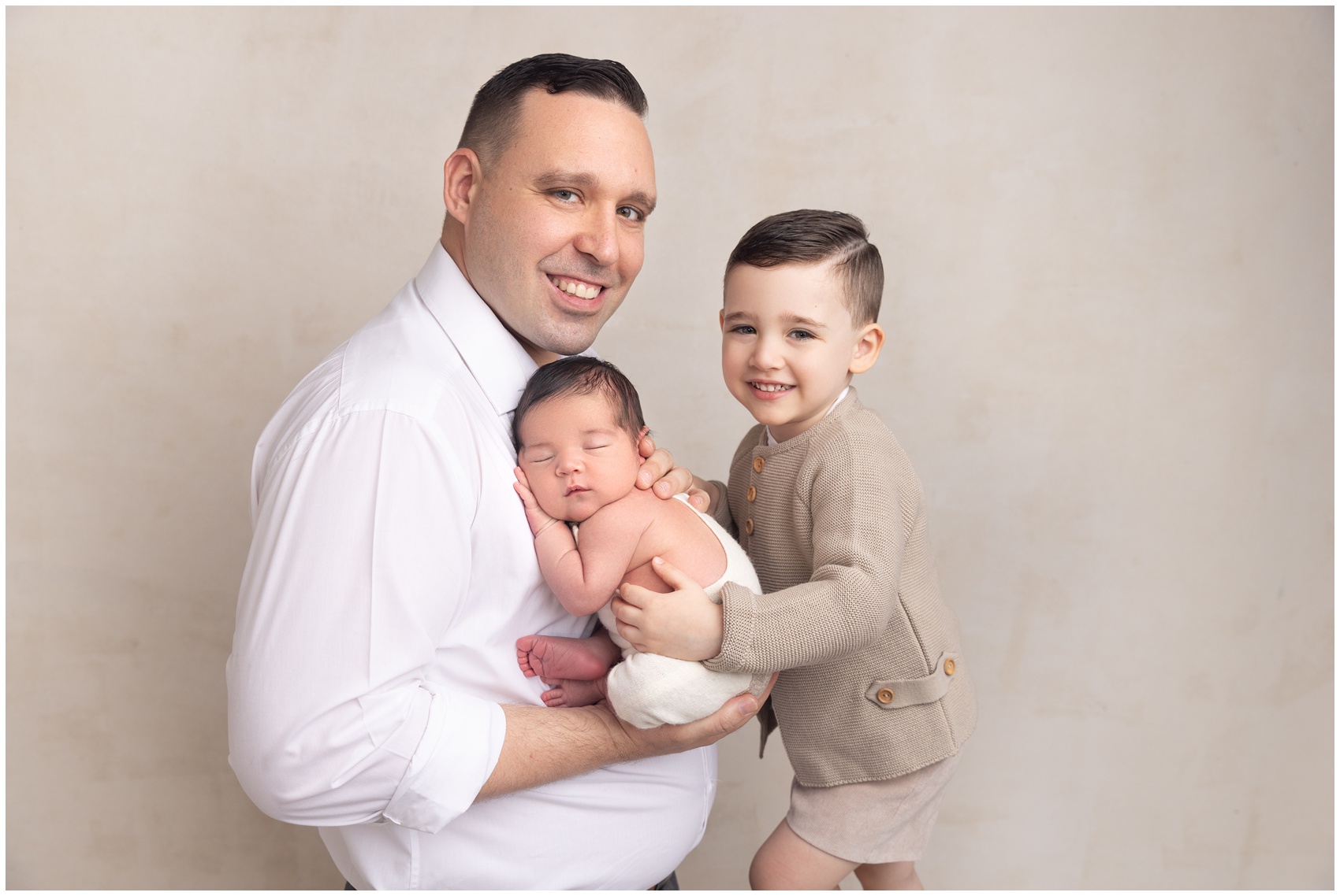 A happy father smiles while standing in a studio cradling his sleeping newborn with his toddler son after visiting hackensack pediatricians