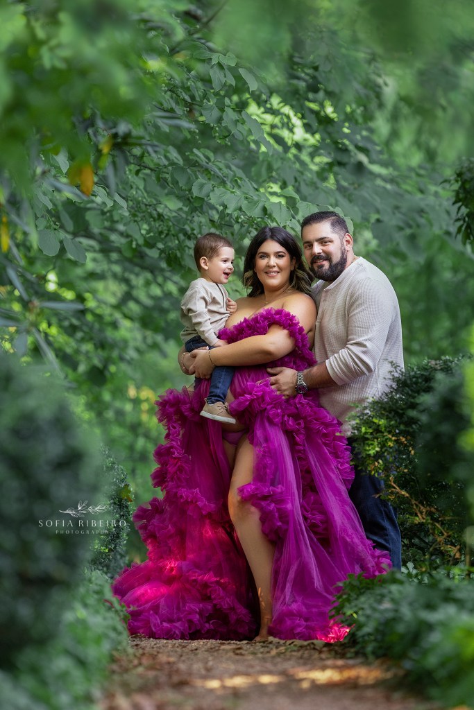 mom and dad smile to the camera while big brother sits atop moms pregnant belly looking at them together