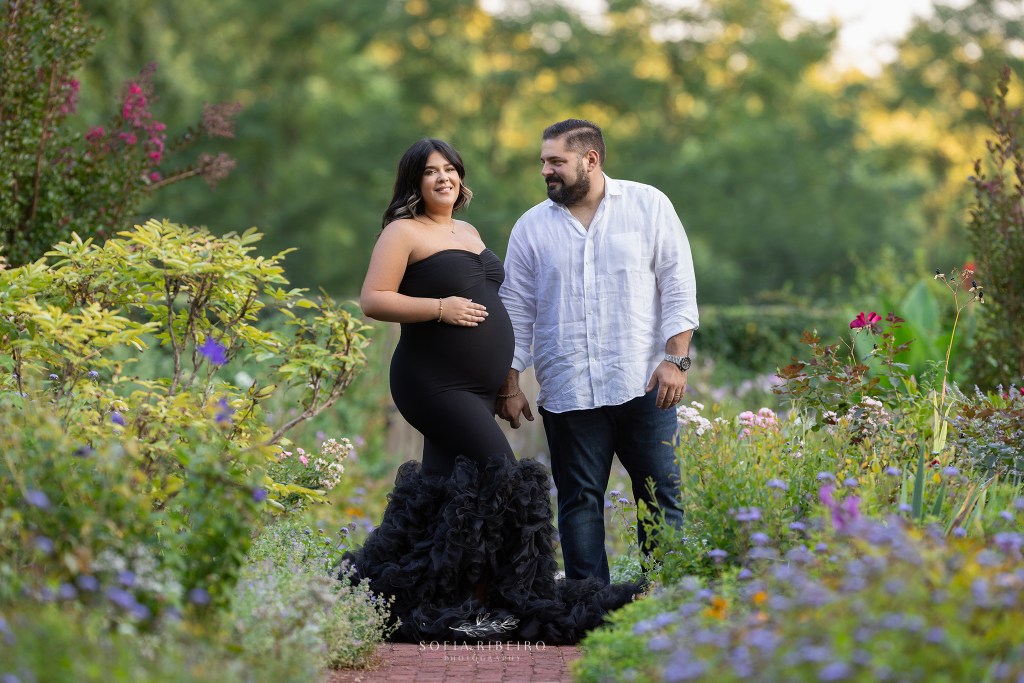 dad looks at his pregnant wife in a black gown, lovingly in a romantic garden in hanover nj