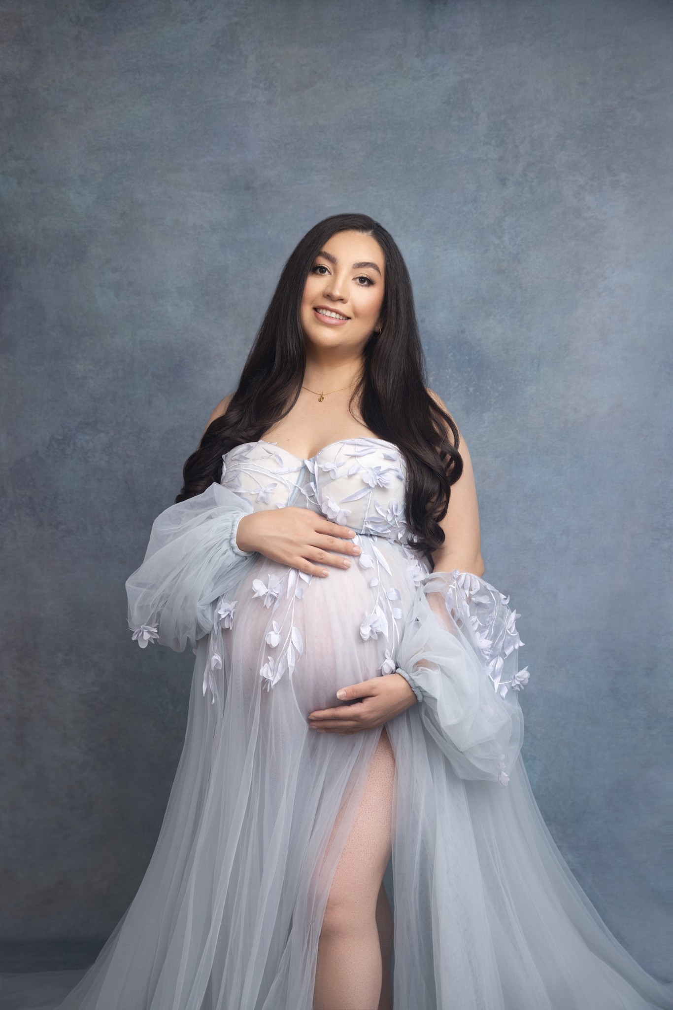 A pregnant woman in a blue maternity gown holds the bump while smiling and standing in a studio after visiting peek-a-boo-baby ultrasound