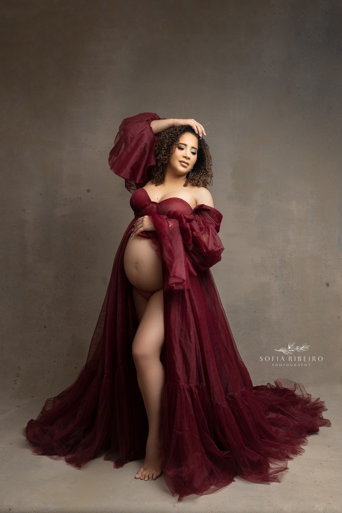 mom poses with exposed belly in a dark red tulle gown, facing the light against a taupe backdrop for maternity portraits