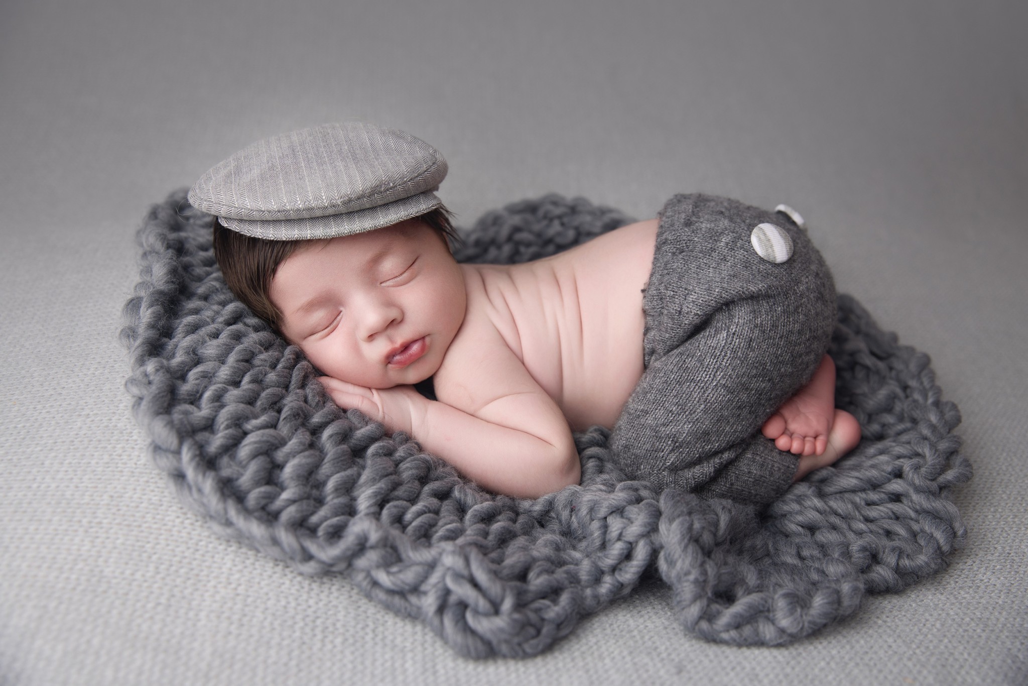 A newborn baby in a grey hat and knit pants sleeps on a matching woven blanket in froggy pose after visiting swaddles baby teaneck nj