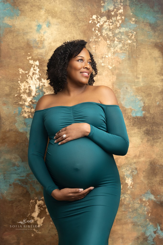 mom smiles into the light in a form fitting teal gown, showcasing her pregnant belly