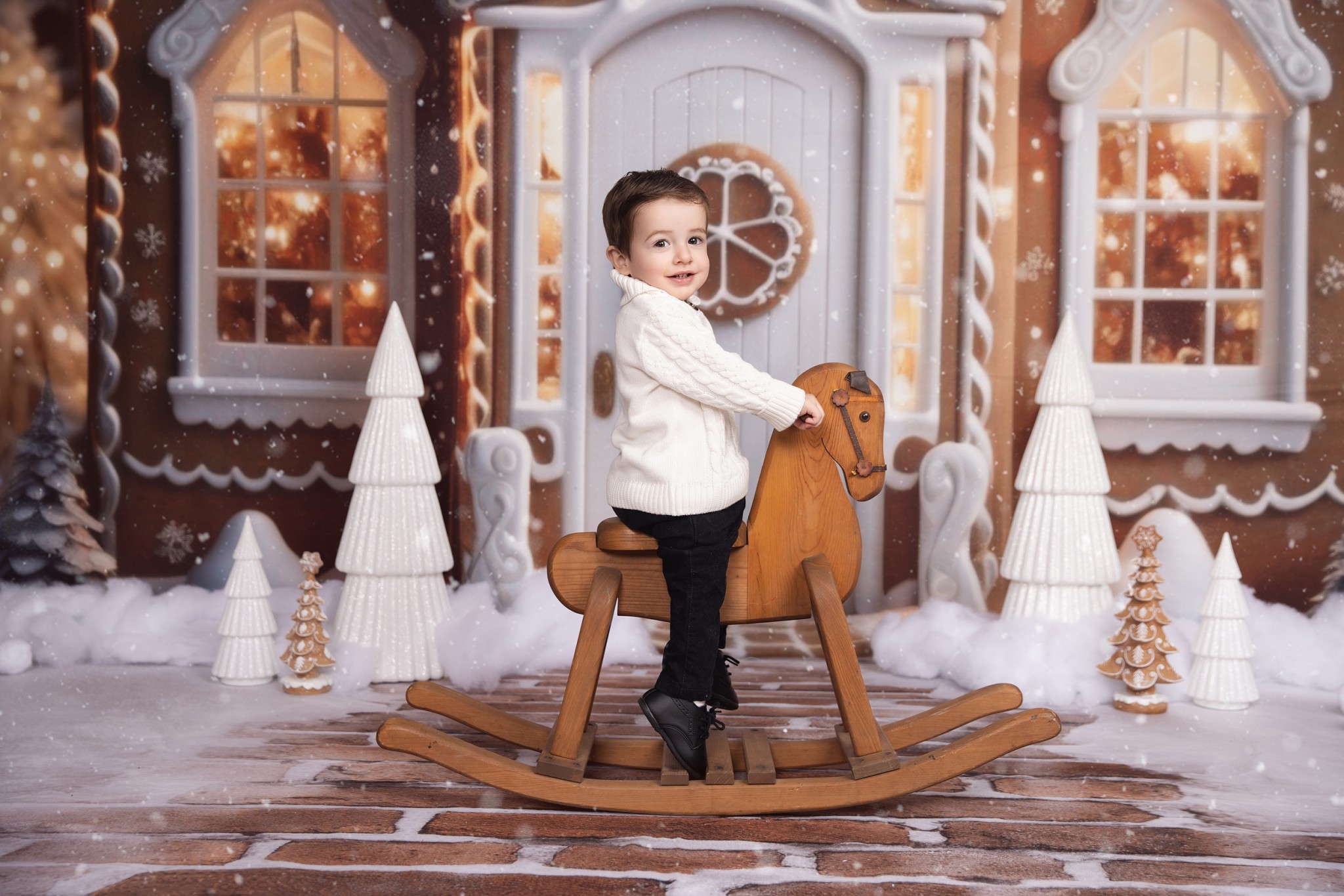 A happy toddler boy in a white sweater sits on a rocking horse in front of a ginger bread house in a photo studio before visiting christmas villages in nj