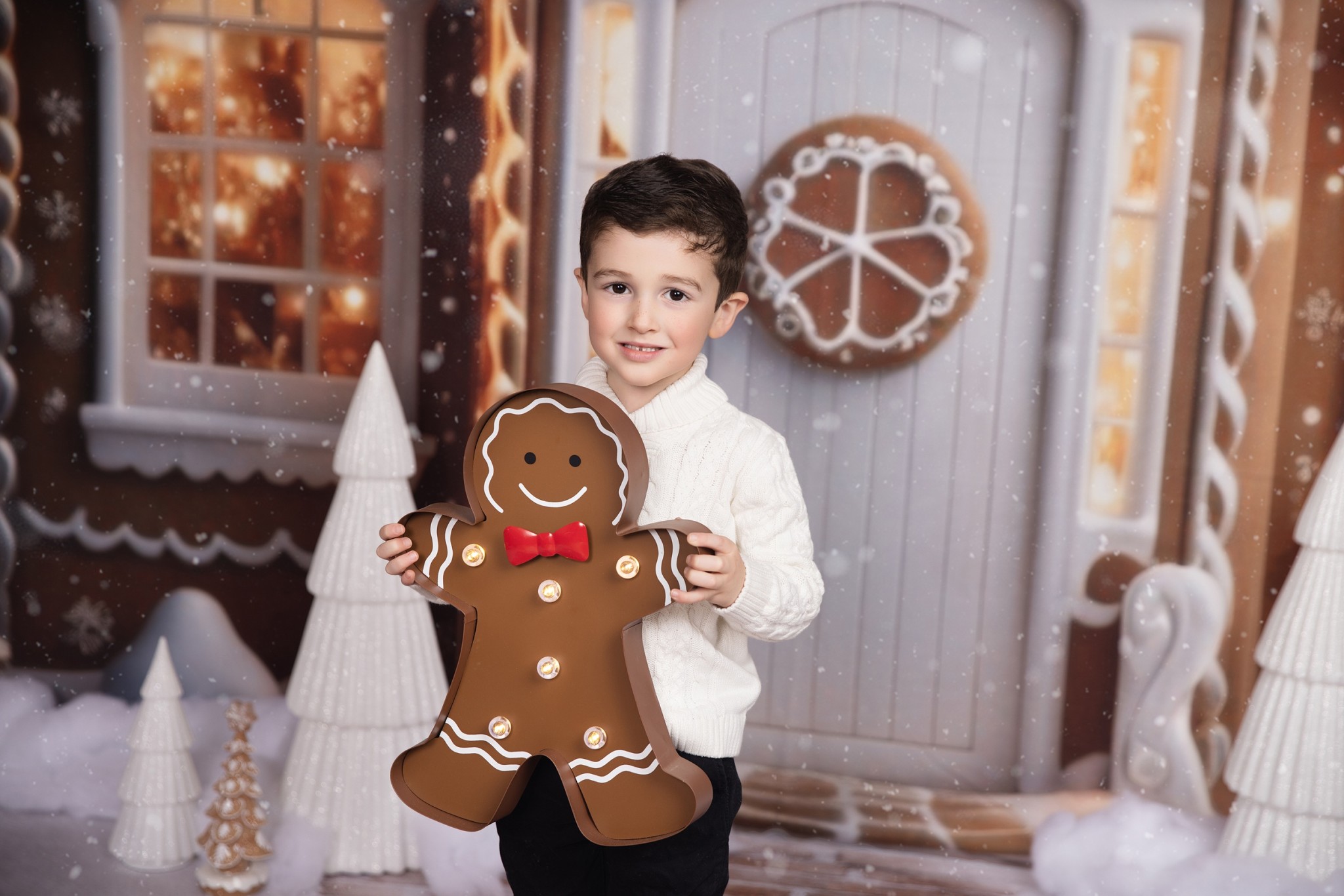 A young boy in a white sweater and black pants stands in a gingerbread house studio holding a light up ginger bread man