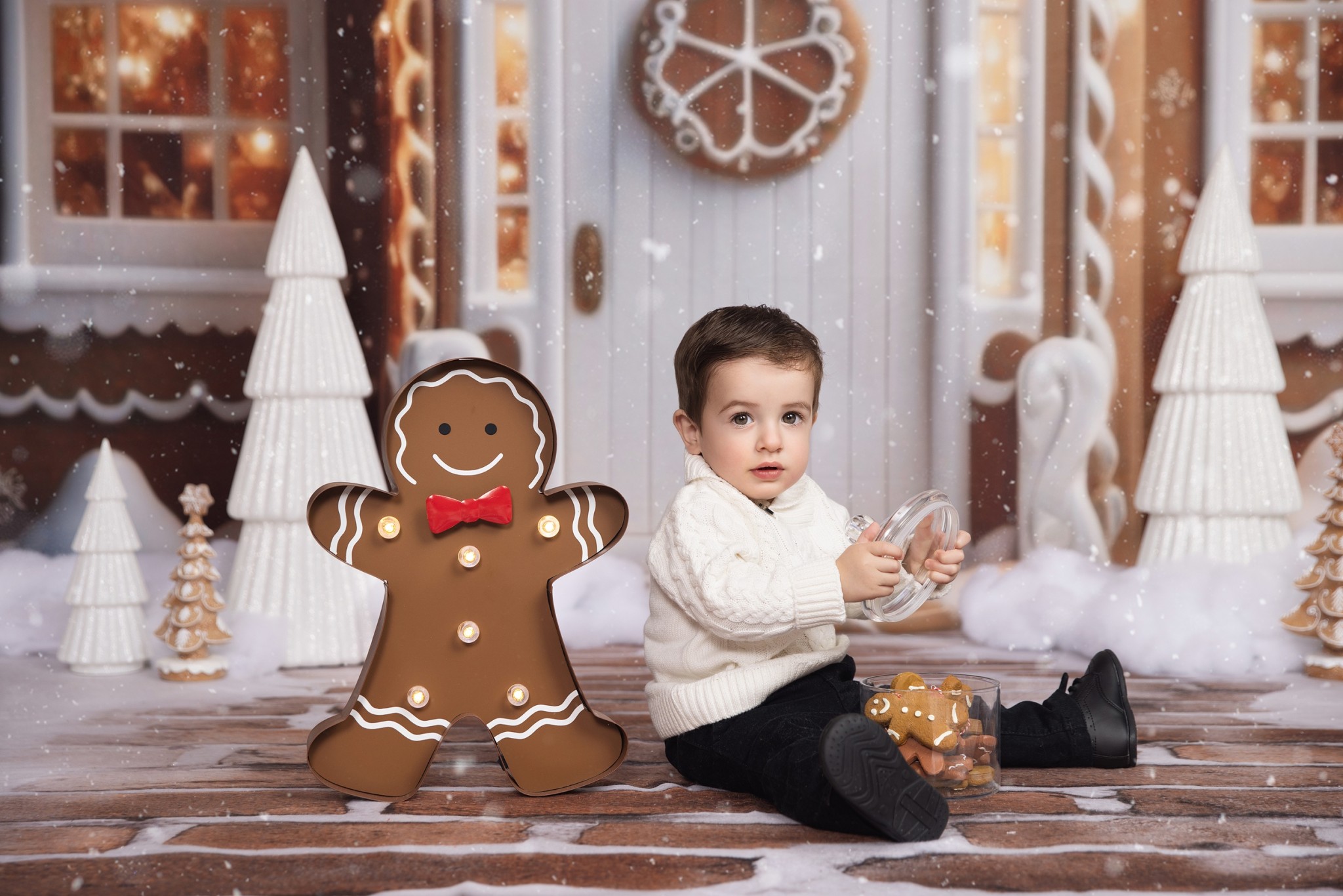 A young toddler sits on a brick path in front of a gingerbread house in a studio opening a jar of gingerbread cookies before visiting christmas villages in nj