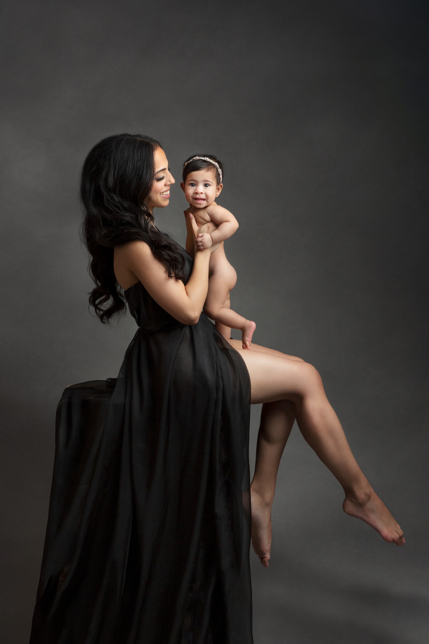A happy baby smiles while being held by mom in a black gown in a studio sitting on a box after visiting daycares in madison nj