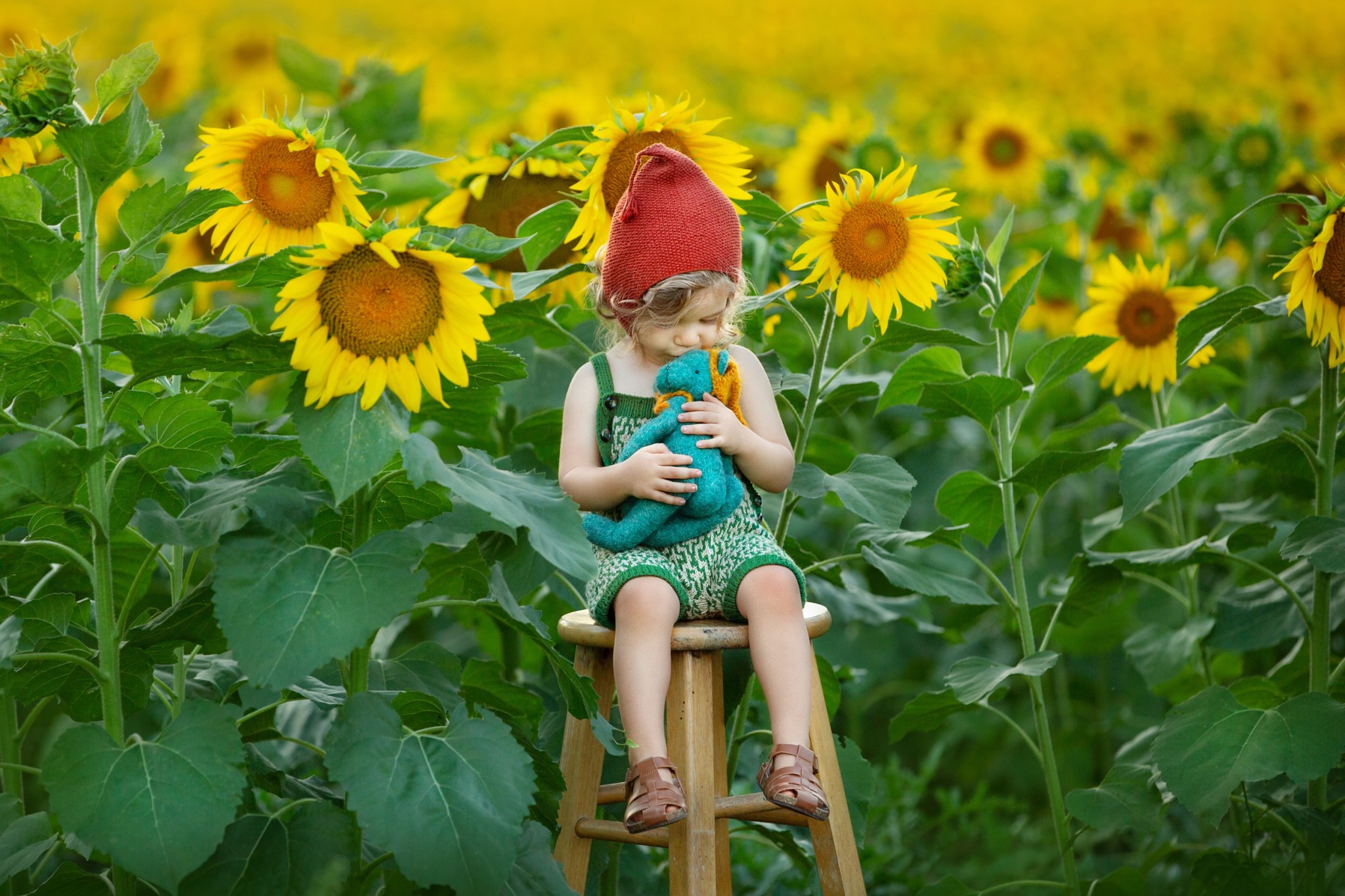 A happy toddler sits on a wooden stool in a field of sunflowers kissing a stuffed toy in a red cap and green knit overalls before visiting halloween events in nj