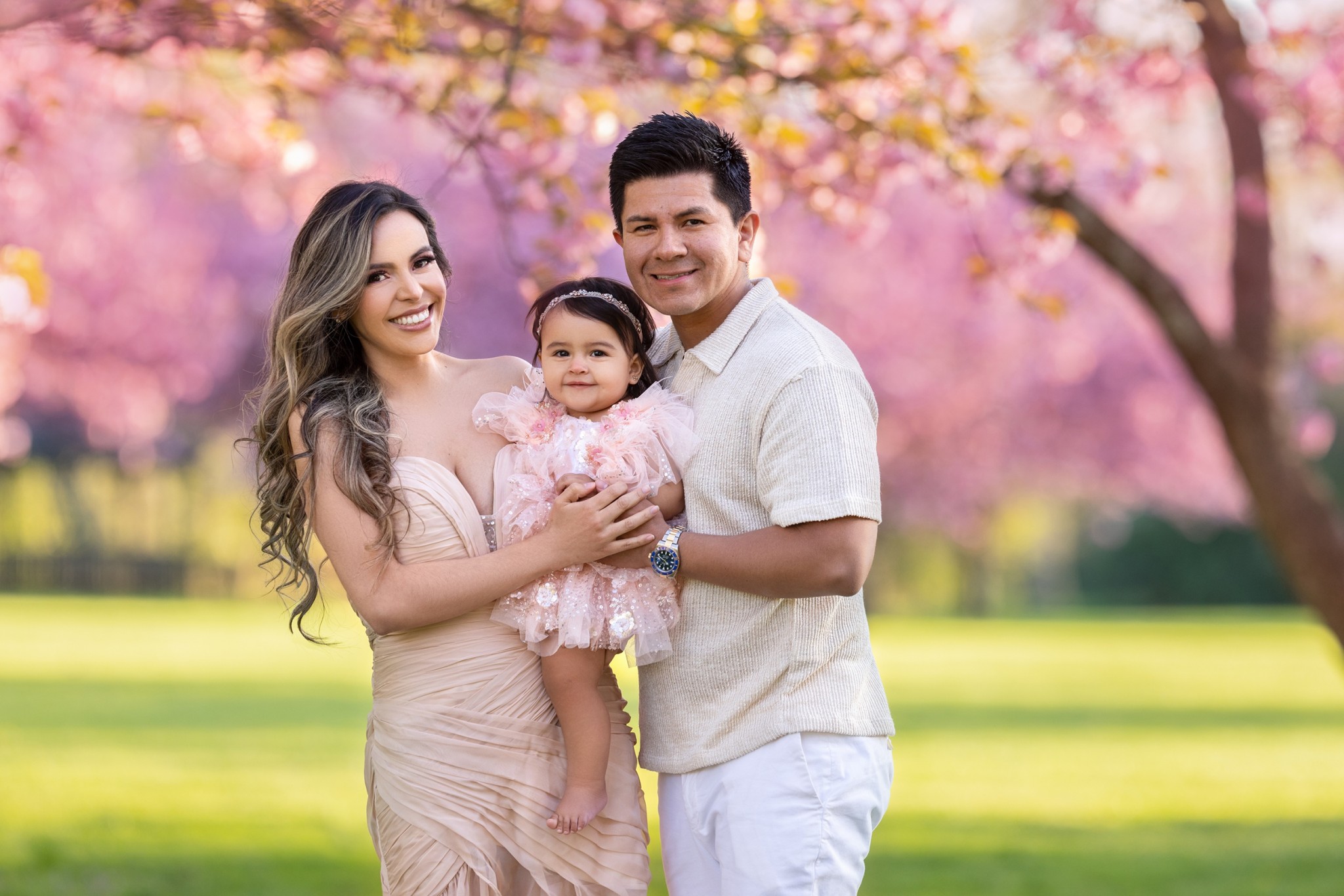Happy mom and dad stand in a park under pink blooming trees holding their baby daughter in a pink dress between them