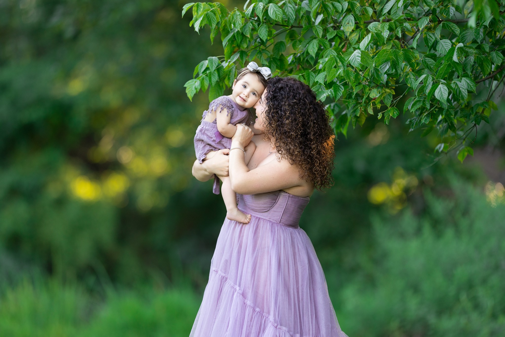 A happy mom in a purple dress stands under a tree playing with her toddler daughter in a matching dress during things to do with hoboken kids