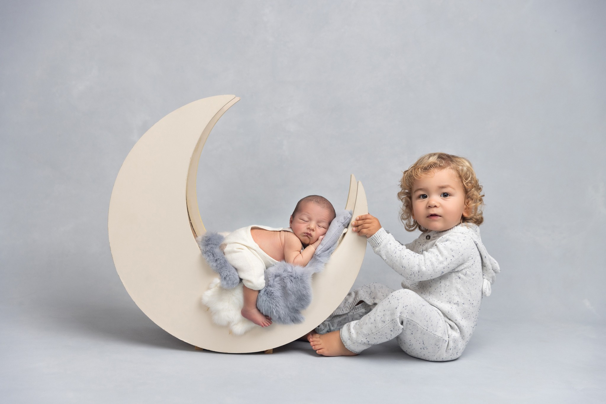A newborn baby sleeps in a moon shaped crib with his older sibling playing in white pajamas with the crib after visiting ladida lakewood