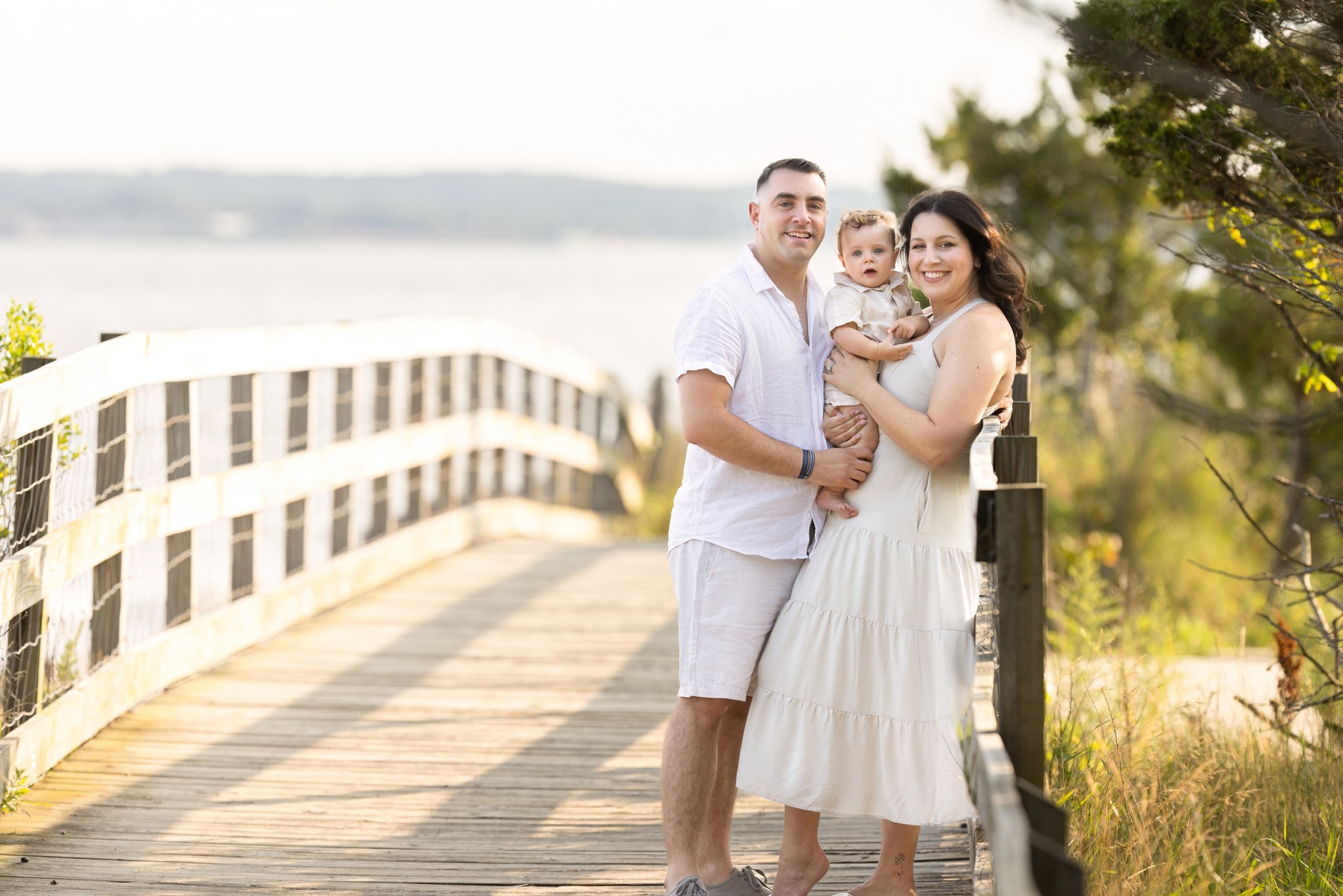 Happy parents lean on a wooden bridge railing while holding their happy baby between them at sunset after some parenting classes jersey city