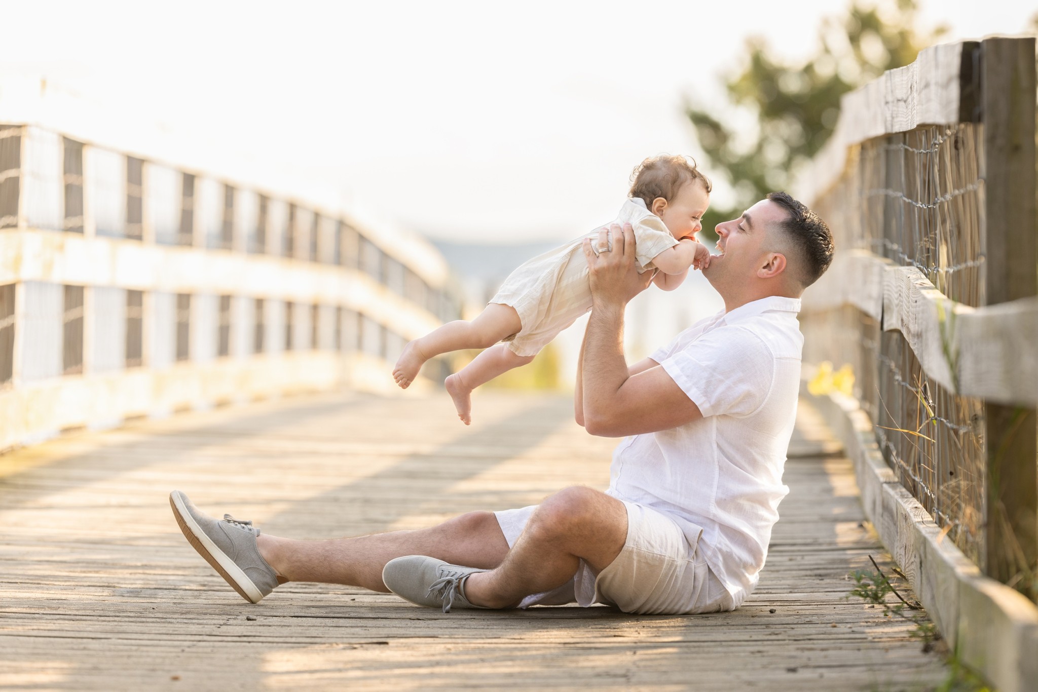 A laughing dad sits on a wooden bridge playing with his infant in white onesie at sunset