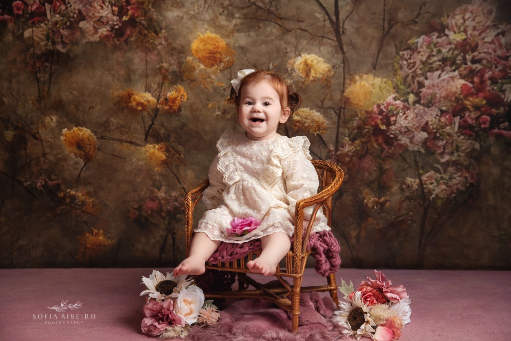 a one year old girl smiles joyfullr in a wicker chair prop against a vintage floral backdrop