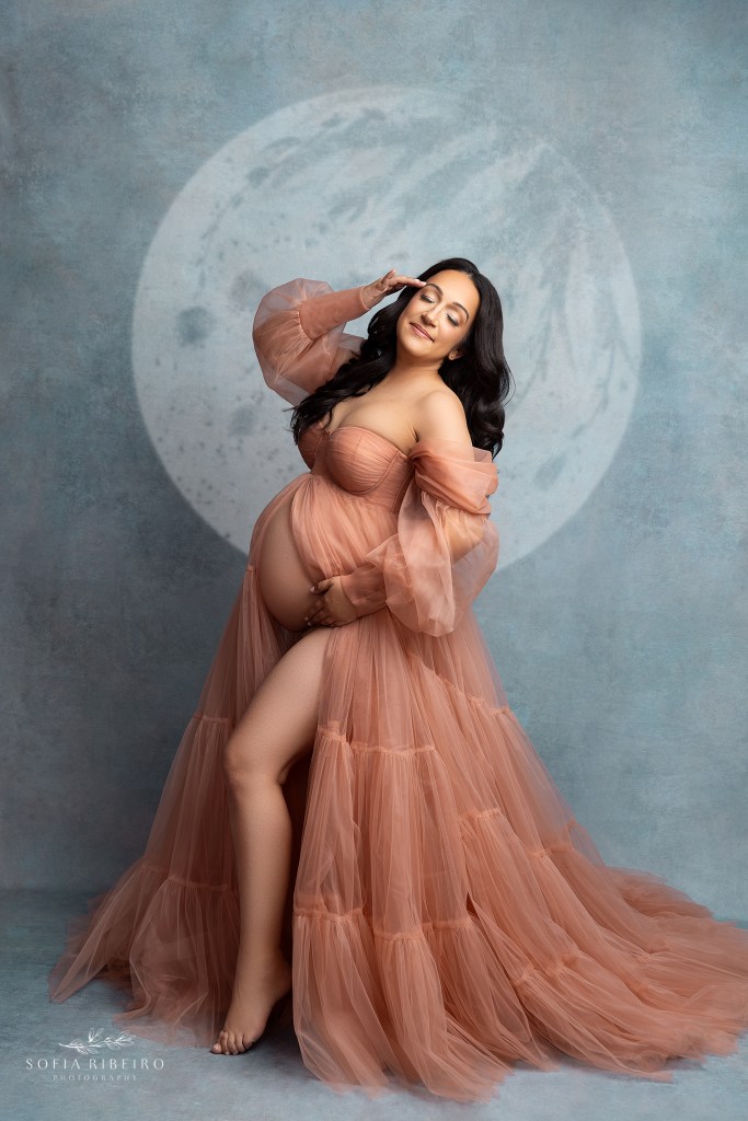 expectant mom poses for maternity portraits with a belleville nj maternity photographer in a peach dress against a blue backdrop