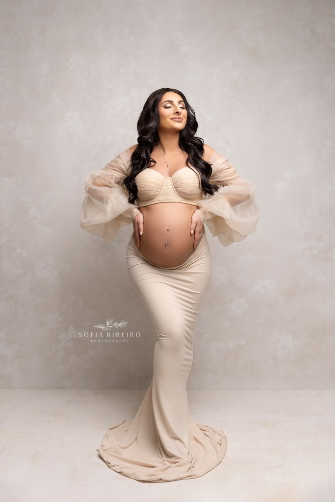 colonia nj maternity photographer poses a pregnant mom in a cream two piece outfit, with hands around the baby bump to emphasize the belly