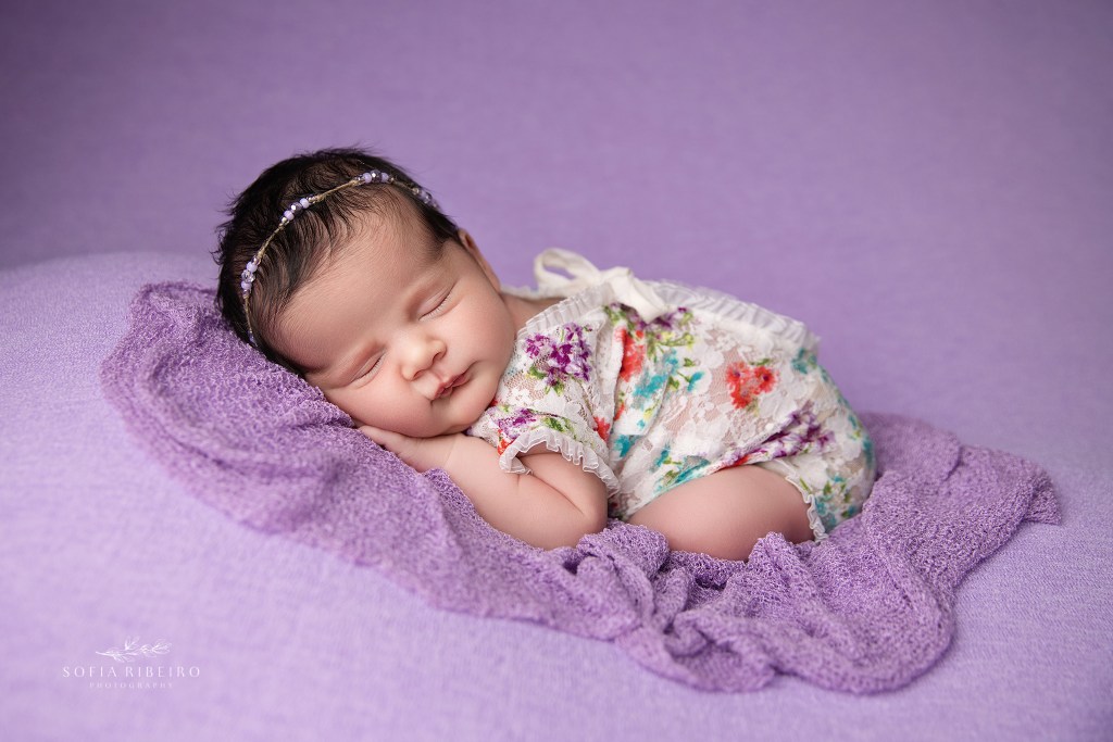 a newborn baby girl is posed on a purple backdrop wearing a white floral outfit