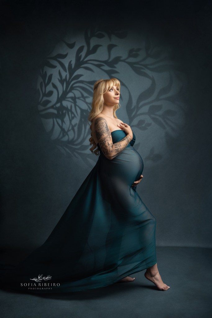 mom is posed against a dark teal backdrop with floral overlay draped in green fabric for maternity photos