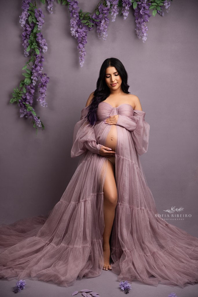 photographer poses an expectant mom in a lavender tulle dress against a matching purple backdrop with flowers