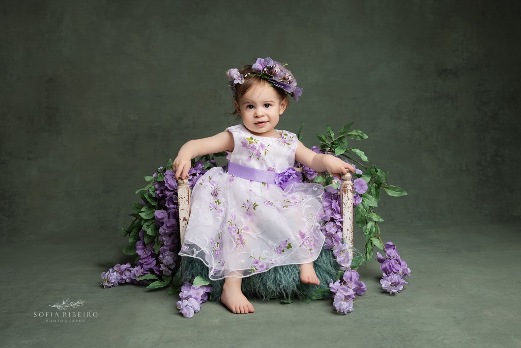 a sweet baby girl poses in a purple and white dress against a green backdrop